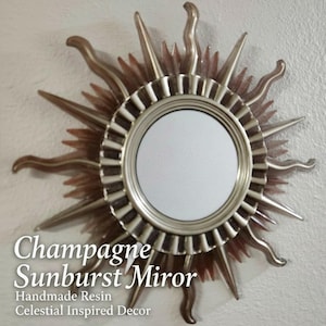 May include: A circular sunburst mirror with a champagne-colored finish. The frame features alternating long, wavy rays and shorter, straight rays surrounding a central circular glass mirror. Text on the image reads: Champagne Sunburst Miror.