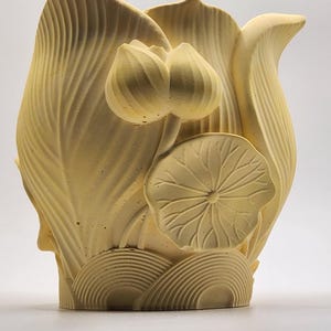 May include: A light yellow, sculpted vase in the shape of a lotus flower. The vase features detailed leaf and petal designs, with a pair of unopened lotus buds and a single, open leaf. The base has a series of curved lines.