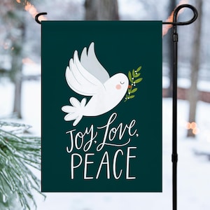 May include: A green garden flag with a white dove carrying a sprig of green leaves. The text "Joy, Love, Peace" is written in white cursive lettering.
