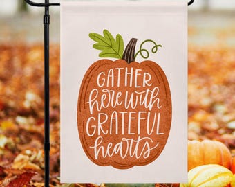 May include: A white garden flag with a brown pumpkin and the text "Gather here with grateful hearts".