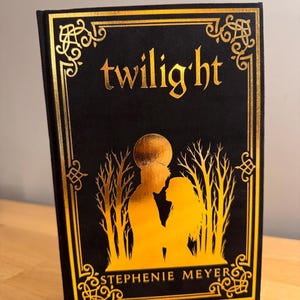 May include: A hardcover book titled "twilight" by Stephenie Meyer. The cover is black with gold foil accents, including the title, decorative borders, and a silhouette of two figures under a moon. The book is displayed on a wooden surface.