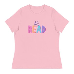 Puede incluir: Una camiseta rosa claro con las palabras "let's READ" en letras de burbujas coloridas. El texto es morado, naranja y azul. La camiseta tiene mangas cortas y cuello redondo.