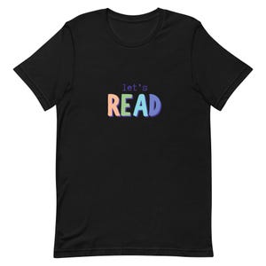 Puede incluir: Camiseta negra con la frase "let's READ" en letras coloridas. La palabra "READ" está en tonos naranja, verde y azul. Una camiseta casual para los amantes de la lectura, perfecta para el uso diario.