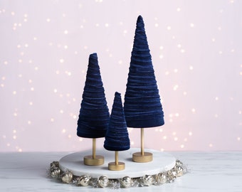 May include: Three blue velvet Christmas trees of varying sizes on a white marble stand with a silver and gold garland around the base.