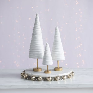 Puede incluir: Tres árboles de Navidad de cerámica blanca de diferentes tamaños en un soporte dorado con una guirnalda plateada alrededor de la base.