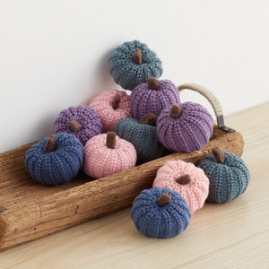 May include: A wooden tray filled with knitted pumpkins in shades of pink, purple, and blue. The pumpkins are arranged in a pile, with some overlapping.