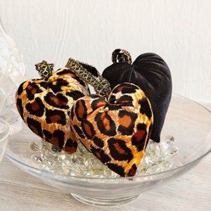 Peut inclure: Trois coussins en forme de cœur dans un bol en verre. Deux des coussins sont recouverts d'un tissu à imprimé léopard et un est en velours noir.