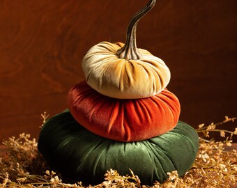 May include: A stack of three velvet pumpkins in green, orange, and beige. The pumpkins are arranged in a pyramid shape and are surrounded by dried flowers.