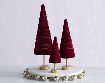 May include: Three burgundy velvet Christmas trees of varying sizes on a gold stand with a silver garland around the base.
