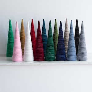 May include: A set of 15 velvet cone Christmas trees in various colors, including pink, white, red, green, blue, and gray. The cones are all the same size and shape, and they are arranged in a row on a white surface.