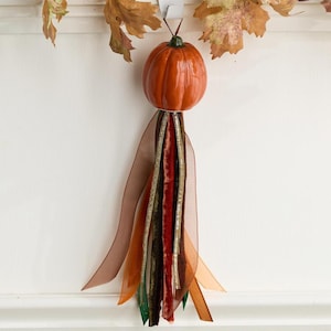 May include: A decorative hanging ornament featuring a glossy orange ceramic pumpkin attached to a white metal hook. Below the pumpkin, a collection of layered ribbons in shades of brown, dark green, and deep red hang down in a tassel style.