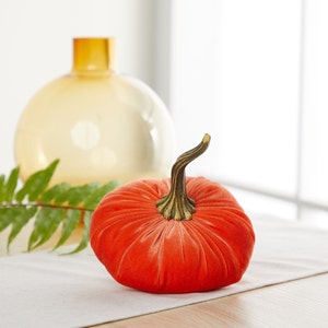 May include: A bright orange velvet pumpkin with a brown stem. The pumpkin is sitting on a white table cloth.
