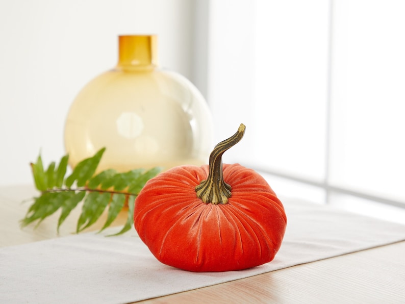 May include: A bright orange velvet pumpkin with a brown stem. The pumpkin is sitting on a white table cloth.