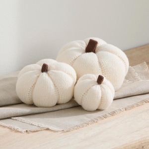 May include: Three ivory-colored, knitted pumpkins of varying sizes are arranged on a beige linen surface. Each pumpkin has a small, brown stem. The pumpkins are a decorative fall or autumn-themed item.