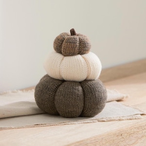 May include: A stack of three knitted pumpkins in varying sizes and colours. The top pumpkin is a mottled brown, the middle pumpkin is a solid cream, and the bottom, largest pumpkin is a solid dark brown. Each pumpkin features a ribbed texture.