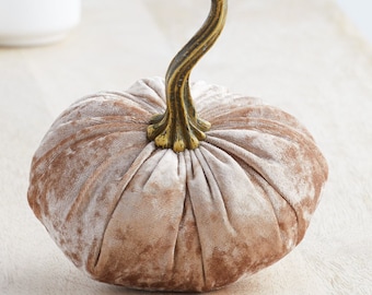 May include: A plush, light brown velvet pumpkin with a bronze-colored, curved stem. The pumpkin has a ribbed texture and is likely a decorative item for fall or autumn-themed home decor.