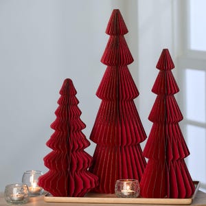 May include: Three red paper Christmas trees of varying sizes are displayed on a wooden tray. The trees have a layered, accordion-like design. Small glass candle holders with lit candles are in front of the trees.