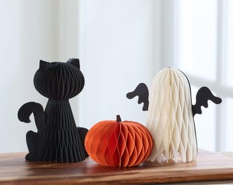 May include: Halloween-themed paper decorations. A black cat, an orange pumpkin, and a white ghost with black wings are displayed on a wooden surface. The decorations are made of folded paper, creating a honeycomb effect.
