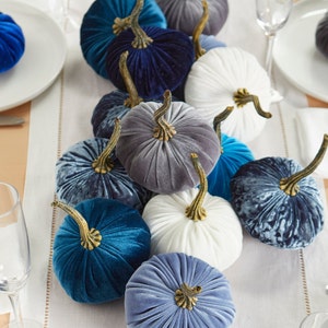 May include: A collection of velvet pumpkins in shades of blue, gray, and white. The pumpkins are arranged on a white tablecloth and are perfect for fall decorating.