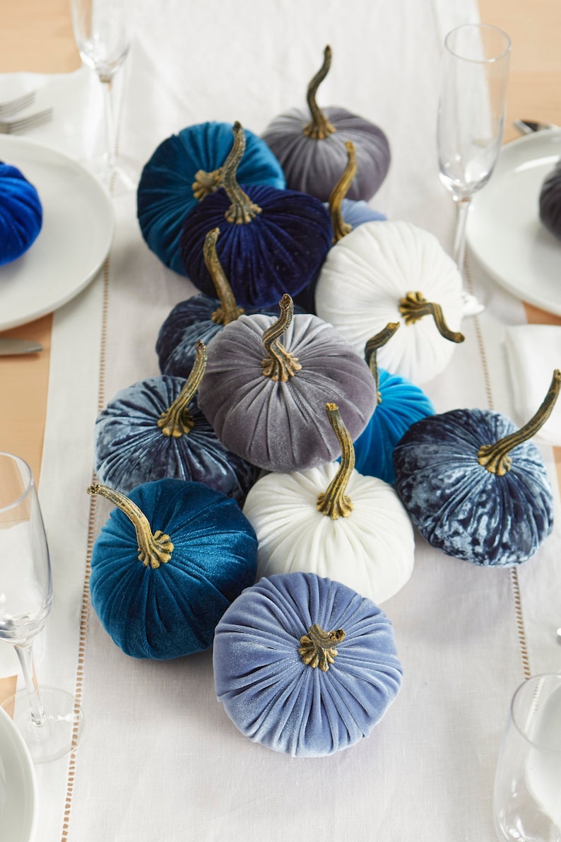 May include: A collection of velvet pumpkins in shades of blue, gray, and white. The pumpkins are arranged on a white tablecloth and are perfect for fall decorating.