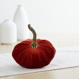 May include: A velvet pumpkin in a deep orange color with a brown stem. The pumpkin is sitting on a white linen table runner.