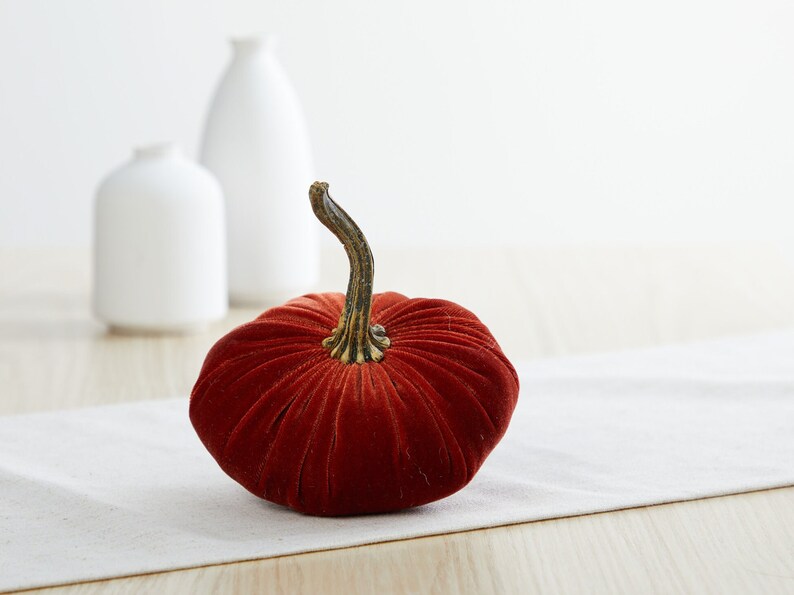 May include: A velvet pumpkin in a deep orange color with a brown stem. The pumpkin is sitting on a white linen table runner.