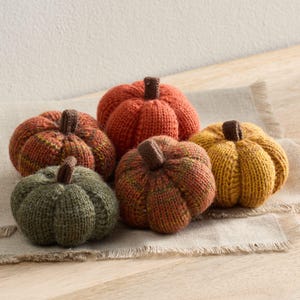 May include: Five knitted pumpkins in autumnal shades of orange, yellow, and green, each with a brown stem. The pumpkins are displayed on a neutral-toned, textured surface, ideal for seasonal decoration. The pumpkins are approximately 10-15 cm in diameter.