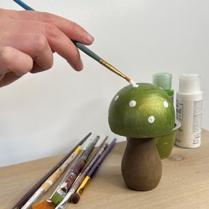 May include: A hand paints white dots on a green mushroom-shaped wooden ornament. The ornament has a brown stem and is sitting on a wooden surface. There are several paintbrushes on the surface.