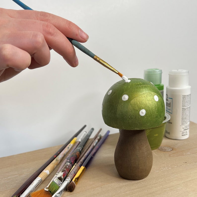 May include: A hand paints white dots on a green mushroom-shaped wooden ornament. The ornament has a brown stem and is sitting on a wooden surface. There are several paintbrushes on the surface.