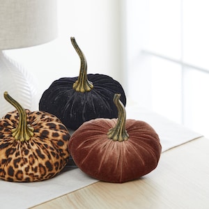 May include: Three velvet pumpkins in different patterns: black, leopard print, and brown. The pumpkins are sitting on a white table runner.