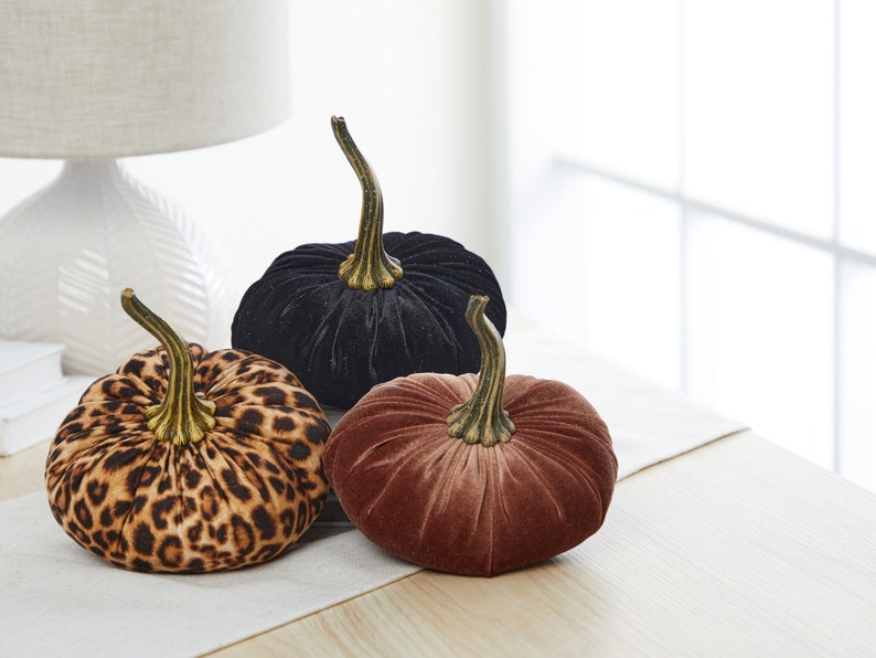 May include: Three velvet pumpkins in different patterns: black, leopard print, and brown. The pumpkins are sitting on a white table runner.