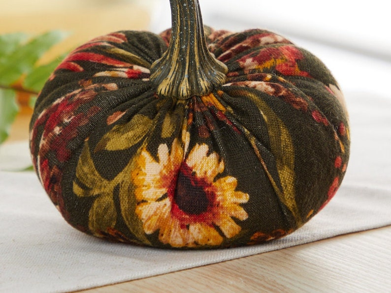 Peut inclure: Une citrouille en tissu vert avec un motif floral compos&eacute; de tournesols, de fleurs rouges et de feuilles vertes. La citrouille a une tige brune.