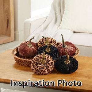 May include: Six velvet pumpkins in a variety of colors and patterns, including leopard print, black, and burgundy. The pumpkins are arranged in a wicker basket on a wooden table.