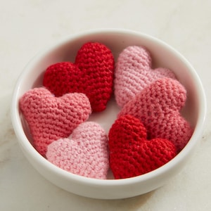 May include: A white bowl filled with six crocheted hearts in shades of pink and red.