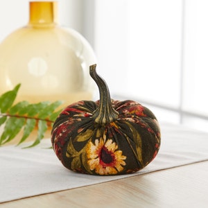 Peut inclure: Une petite citrouille en tissu avec un motif floral dans des tons de vert, rouge et jaune. La citrouille est pos&eacute;e sur une surface blanche.