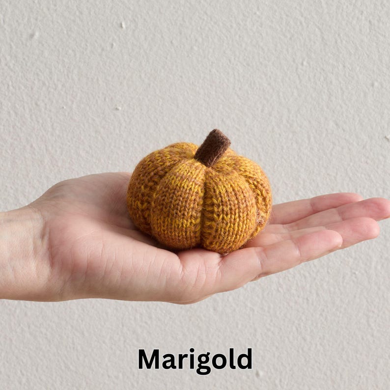 May include: A knitted pumpkin in shades of yellow and brown, with a small brown stem. The pumpkin is held in a hand, and the word "Marigold" is written in black at the bottom. The pumpkin is a decorative item.