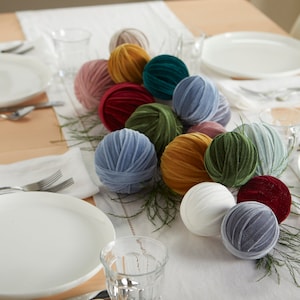May include: A table setting with a centerpiece of velvet balls in various colors, including blue, green, red, and gold. The balls are arranged in a line down the center of the table, with sprigs of greenery interspersed between them.