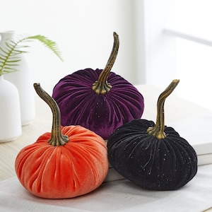 May include: Three velvet pumpkins in orange, purple, and black. The pumpkins are decorated with gold stems and have a ribbed texture.