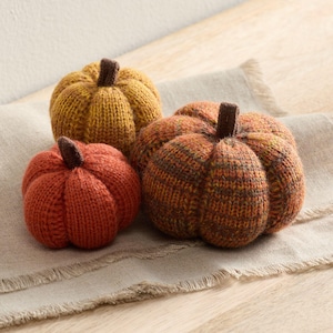 Könnte beinhalten: Drei gestrickte Kürbisse in Orange- und Gelbtönen liegen auf einer neutralfarbenen Leinenoberfläche. Die Kürbisse haben braune Stiele und unterschiedliche Größen, ideal für die Herbstdekoration. Der kleinste Kürbis ist einfarbig orange, während der größte ein gesprenkeltes Muster hat.