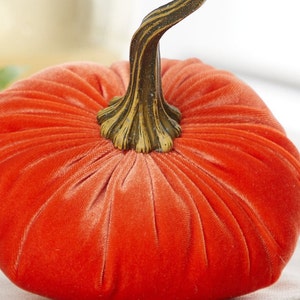May include: A bright orange velvet pumpkin with a brown, textured stem. The pumpkin is sitting on a white surface.