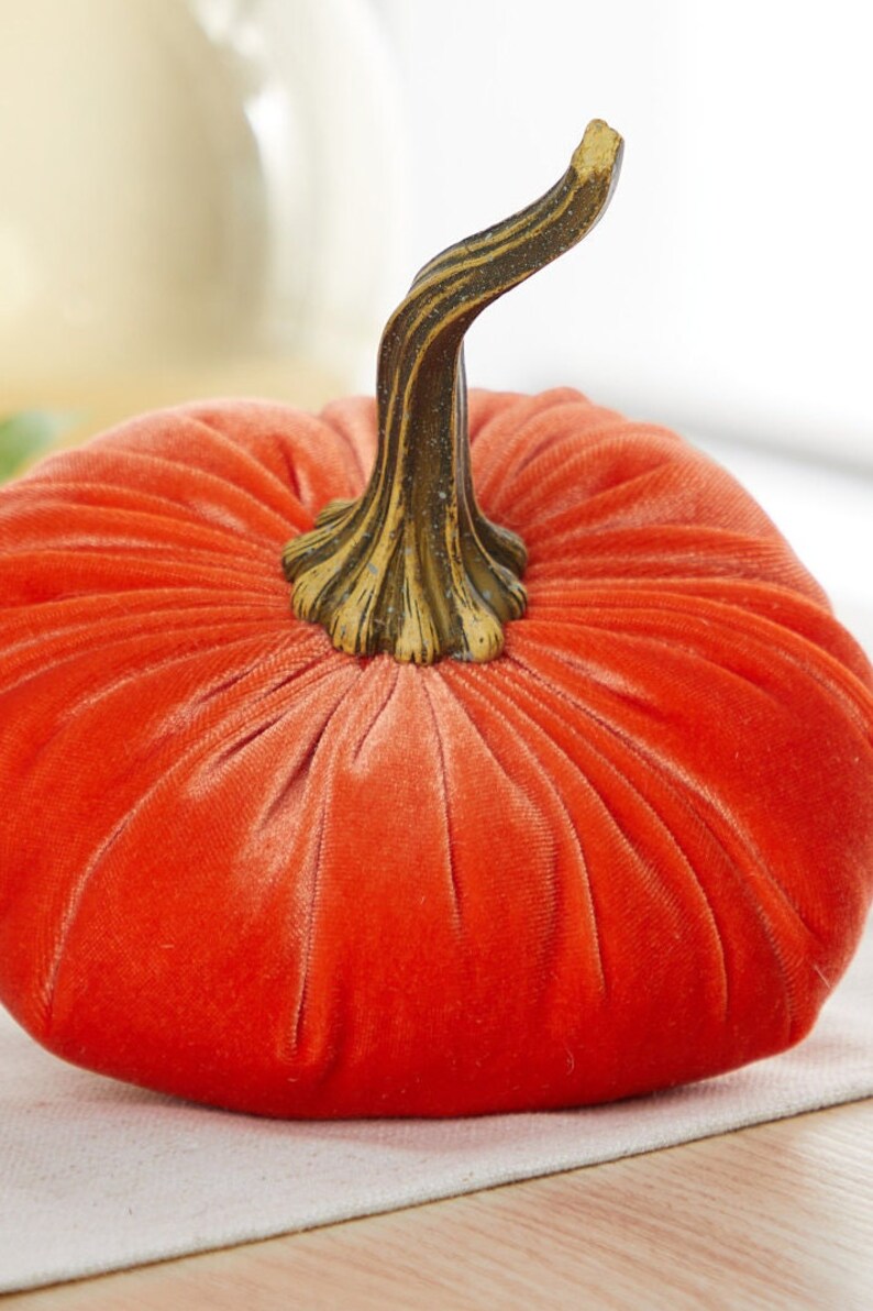 May include: A bright orange velvet pumpkin with a brown, textured stem. The pumpkin is sitting on a white surface.