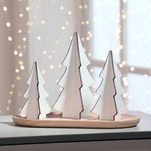 May include: A set of three white wooden Christmas tree decorations with brown edges, displayed on a light brown wooden tray. The trees are different sizes and have a minimalist design, perfect for holiday decor.