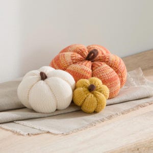 May include: Three knitted pumpkins in varying sizes and colors. One large pumpkin features an orange and yellow variegated yarn pattern, one medium pumpkin is solid off-white, and one small pumpkin is solid mustard yellow. Each has a brown stem.