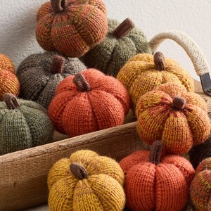 May include: A collection of knitted pumpkins in various autumnal colors, including orange, yellow, and green. Each pumpkin has a brown stem and is arranged in a wooden crate, perfect for fall decor.