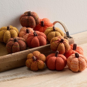 May include: A collection of knitted pumpkins in various shades of orange, brown, and yellow. Each pumpkin has a brown stem and is displayed in a wooden tray, perfect for fall decor.