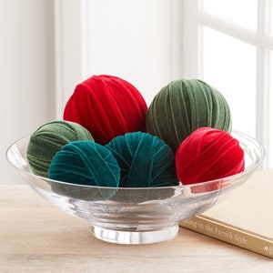 Puede incluir: Un cuenco de vidrio transparente lleno de seis bolas decorativas. Las bolas están hechas de terciopelo rojo y verde y tienen una textura acanalada.