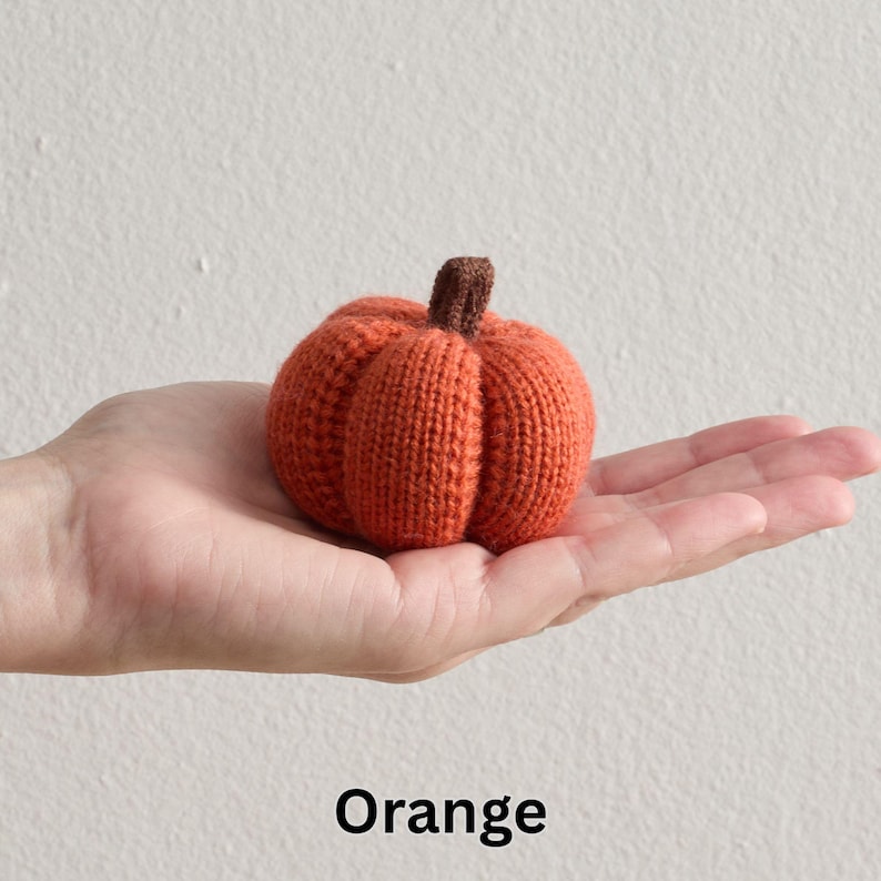 May include: An orange knitted pumpkin with a brown stem. The pumpkin is held in a hand, and the word "Orange" is written in black at the bottom of the image. The pumpkin is a seasonal decoration.
