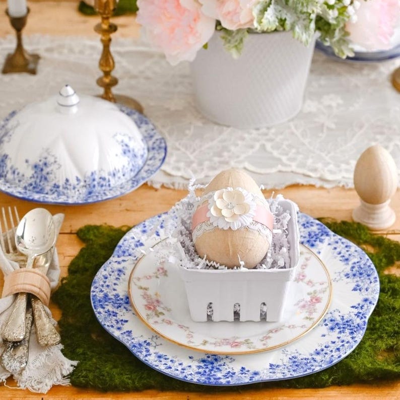 May include: A white ceramic plate with a blue floral pattern, a white ceramic bowl with a blue floral pattern, and a white ceramic egg holder with a pink flower and ribbon. The egg holder is filled with white shredded paper and a white egg with a pink flower and ribbon.