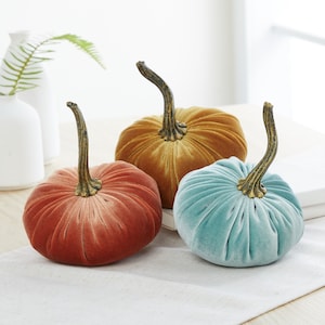 May include: Three velvet pumpkins in different colors: orange, gold, and teal. The pumpkins are all the same size and have a slightly ruffled texture.