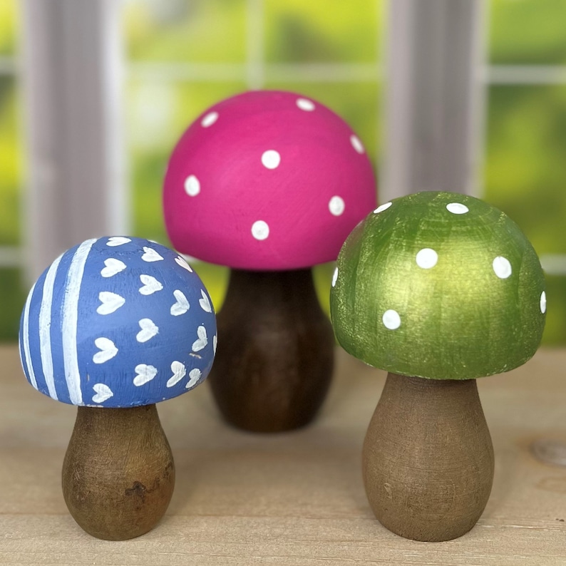 May include: Three wooden mushroom figurines with white polka dots. The mushrooms are painted in pink, green, and blue.